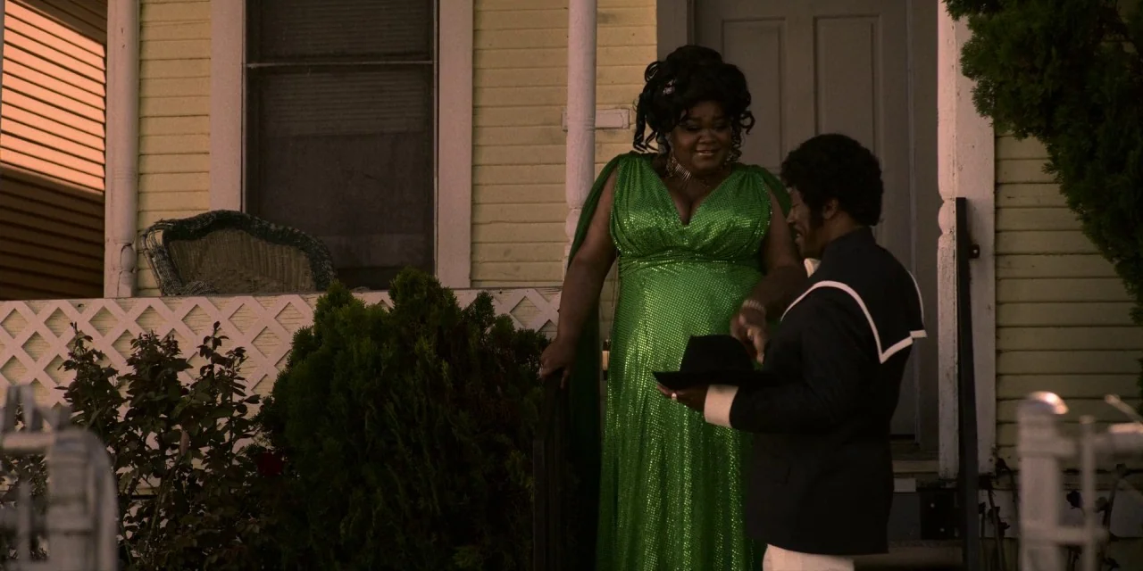 Dolemite Is My Name movie still: medium — A light yellow wooden house porch with a white lattice fence and green bushes. A woman in a sparkly green…