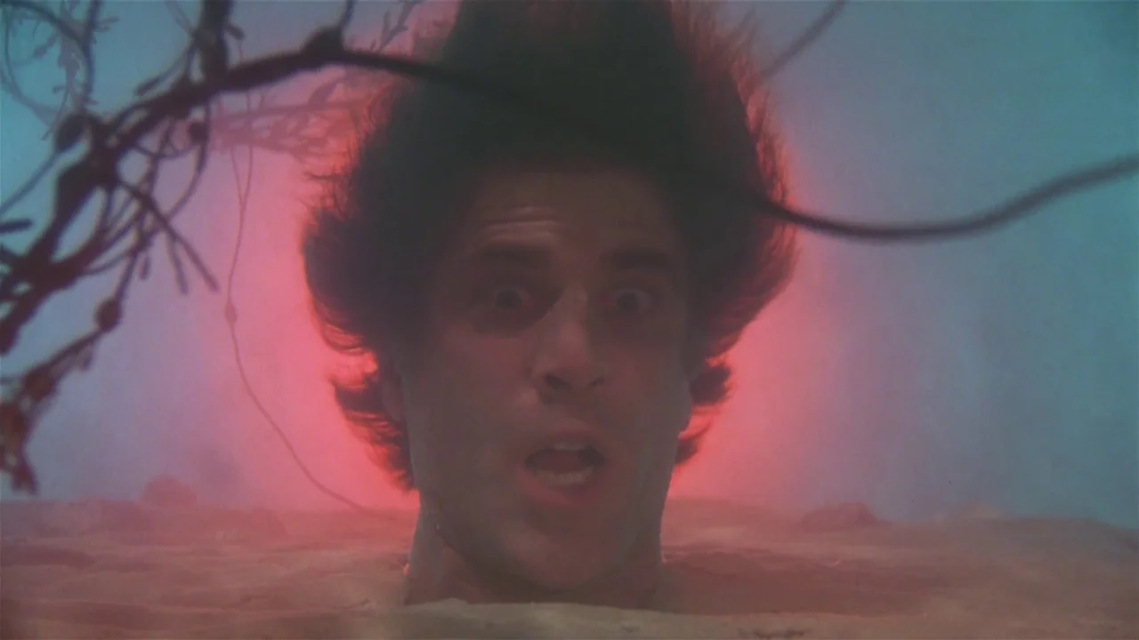 Creepshow (1982) movie still: closeup — A man's head protrudes from the sand at night in a desolate desert setting. A glowing red light emanates…