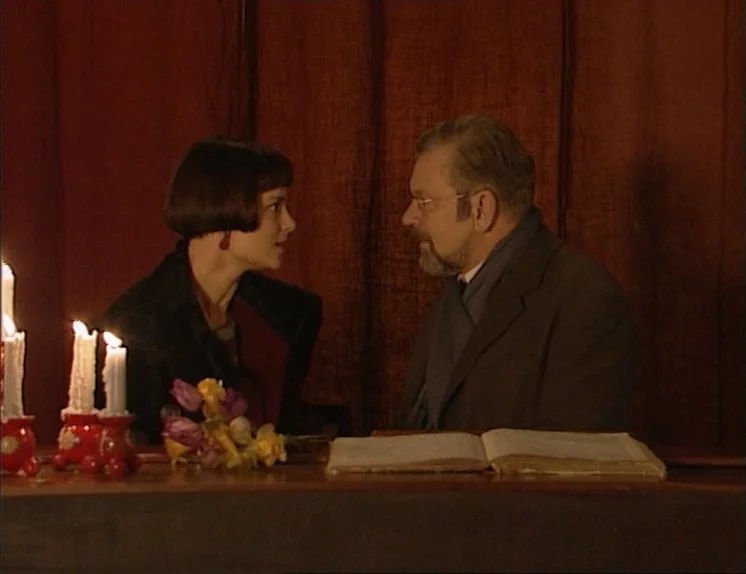 In The Presence of a Clown (1997) movie still: medium — A dimly lit room features a table set with lit candles and an open book. A woman with a dark…