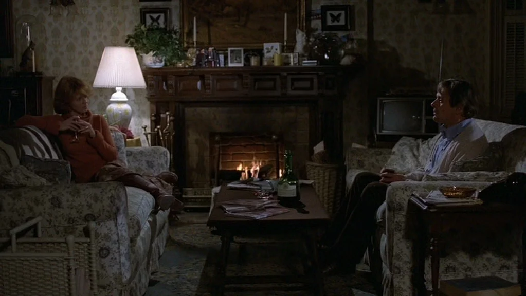 Magic (1978) movie still: wide — A dimly lit living room features floral patterned sofas and a brick fireplace. A man and a woman sit facing each…