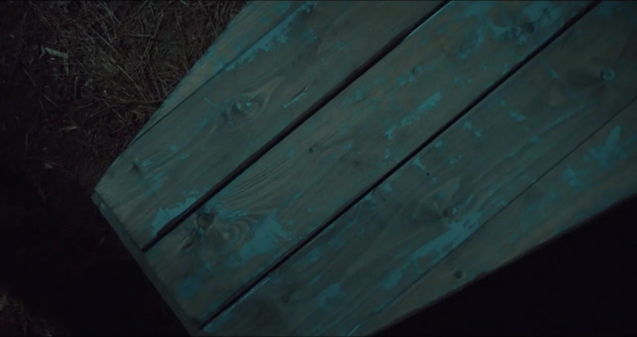 Eye for an Eye (2025) movie still: closeup — A close-up of a weathered wooden coffin lid resting on dark forest soil. The surface shows peeling blue…