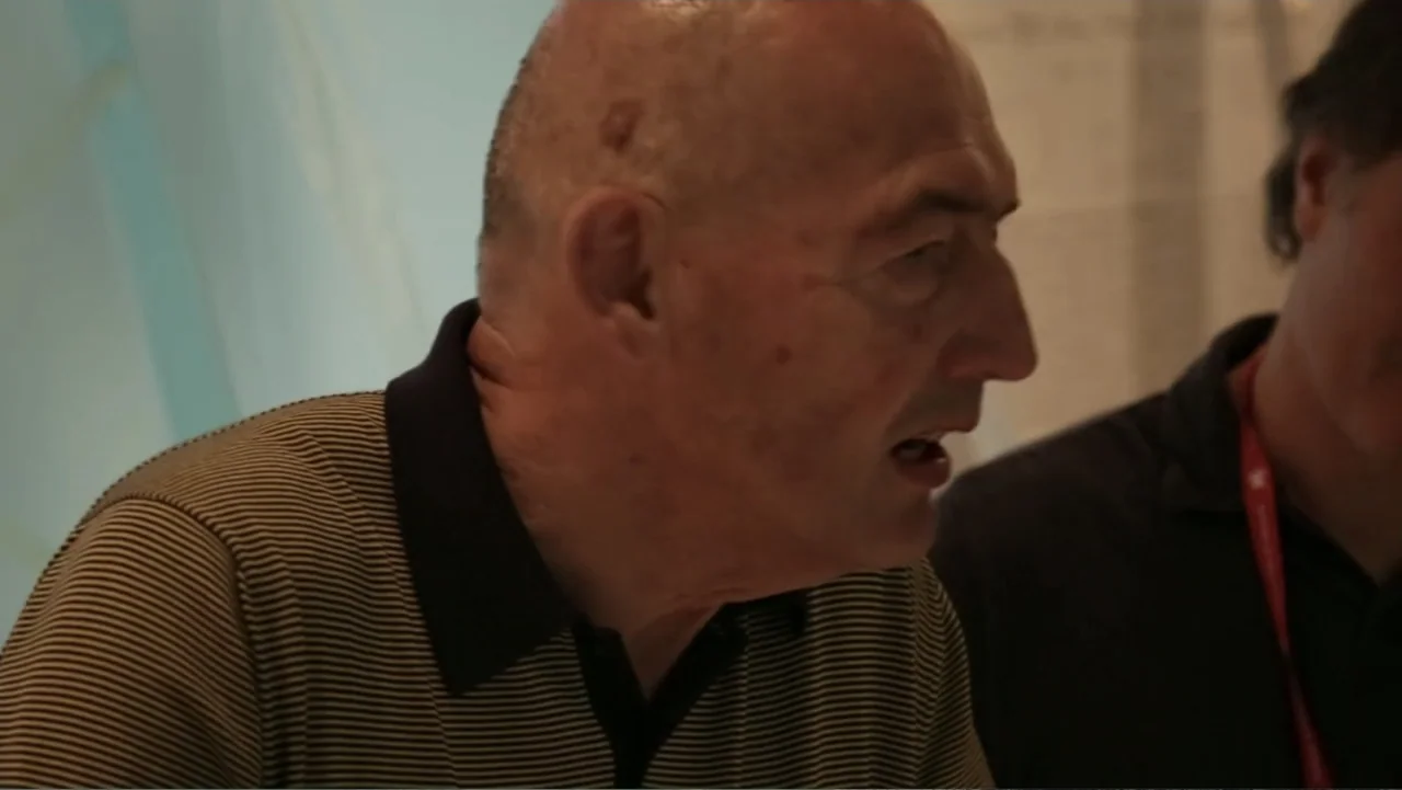 REM (2019) movie still: closeup — A room with textured walls hosts two men in a close-up conversation. An older man in a striped shirt speaks while…