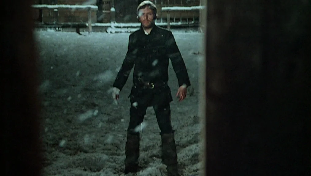 The Great Silence (1968) movie still: fullbody — A snowy outdoor ground at night with dark structures visible in the distance. A man stands alone in…