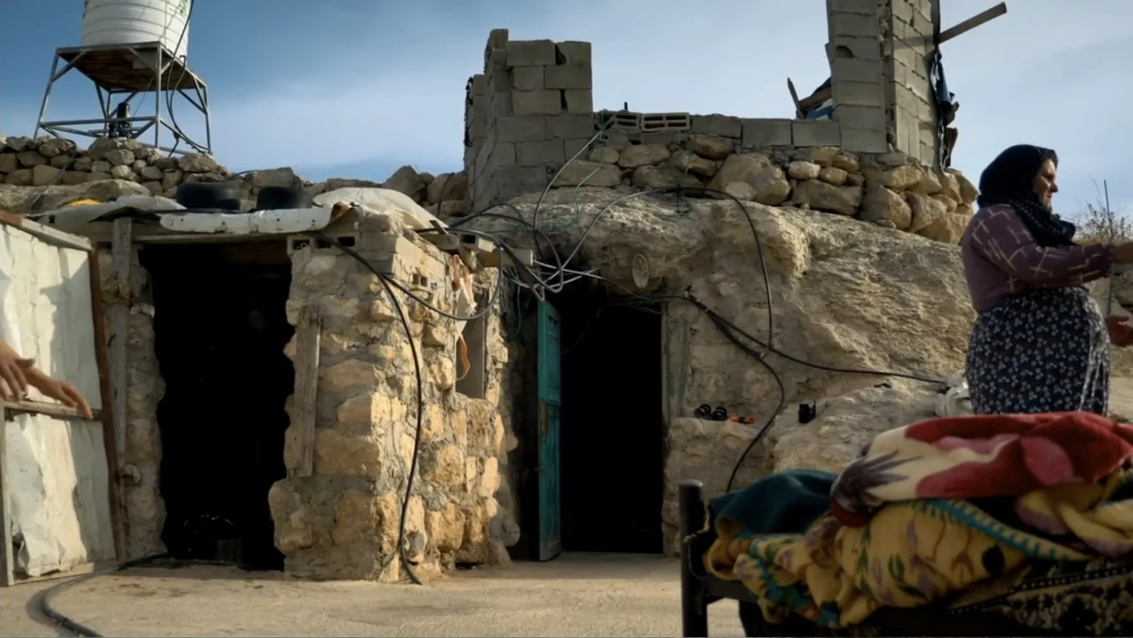 No Other Land (2024) movie still: medium — A rural stone dwelling is built into a rocky hillside under a clear sky. A woman in a headscarf stands to…