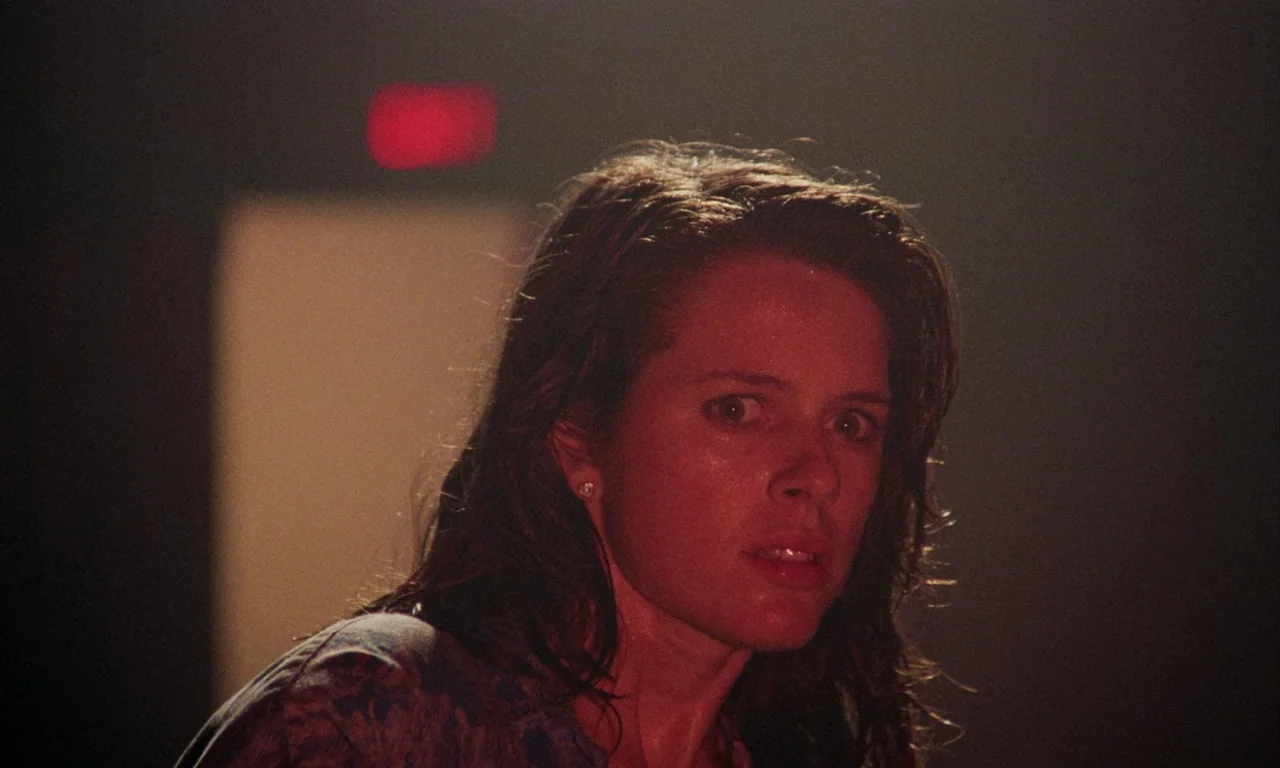 Demons (1985) movie still: closeup — A dark, dimly lit corridor features an out-of-focus red exit sign in the background. A woman with wet hair…