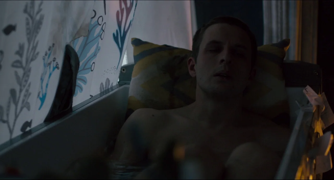 The Night Eats the World (2018) movie still: medium — A man lies shirtless in a bathtub with a patterned shower curtain pulled partially closed. The…
