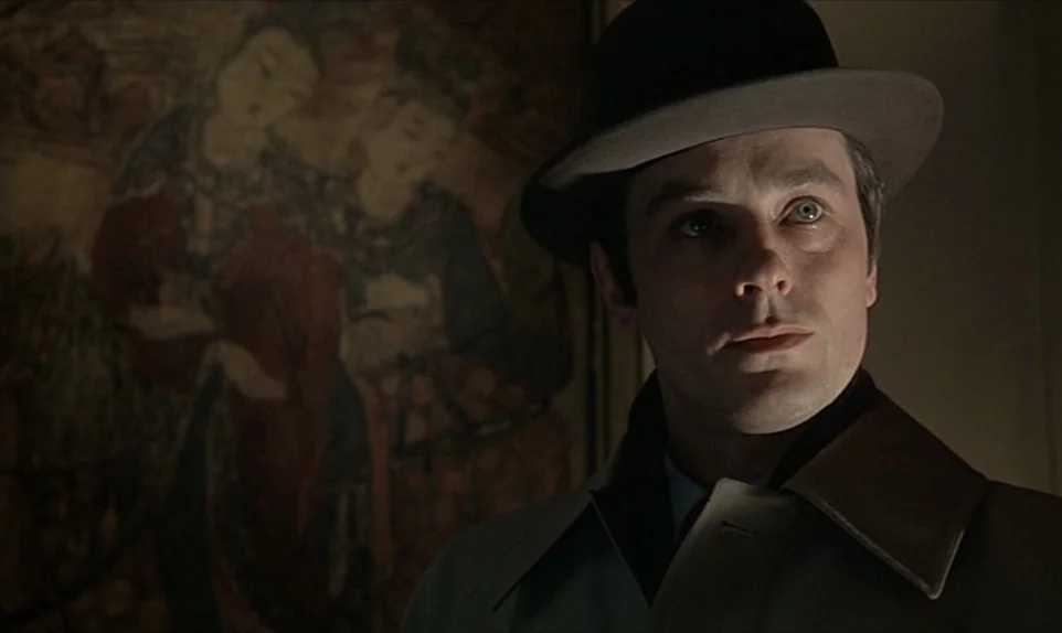Mr. Klein (1976) movie still: medium — A dimly lit room features a large, dark painting of two figures on the wall. A man in a hat and trench coat…