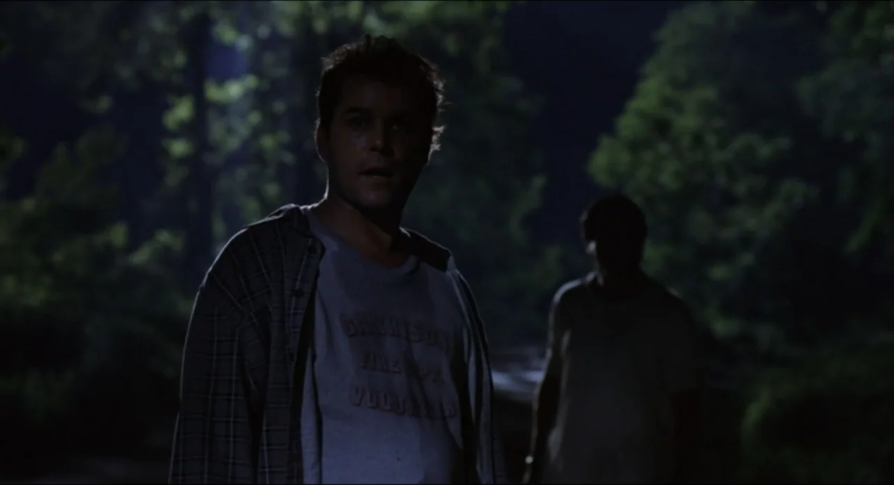 Cop Land (1997) movie still: medium — A dark, wooded area is dimly lit by light during the night. A man in a t-shirt looks forward while another…