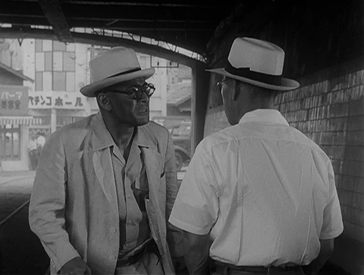 I Live In Fear (1955) movie still: medium — Two men in fedoras stand under a covered walkway on a city street. A Japanese business sign is visible…