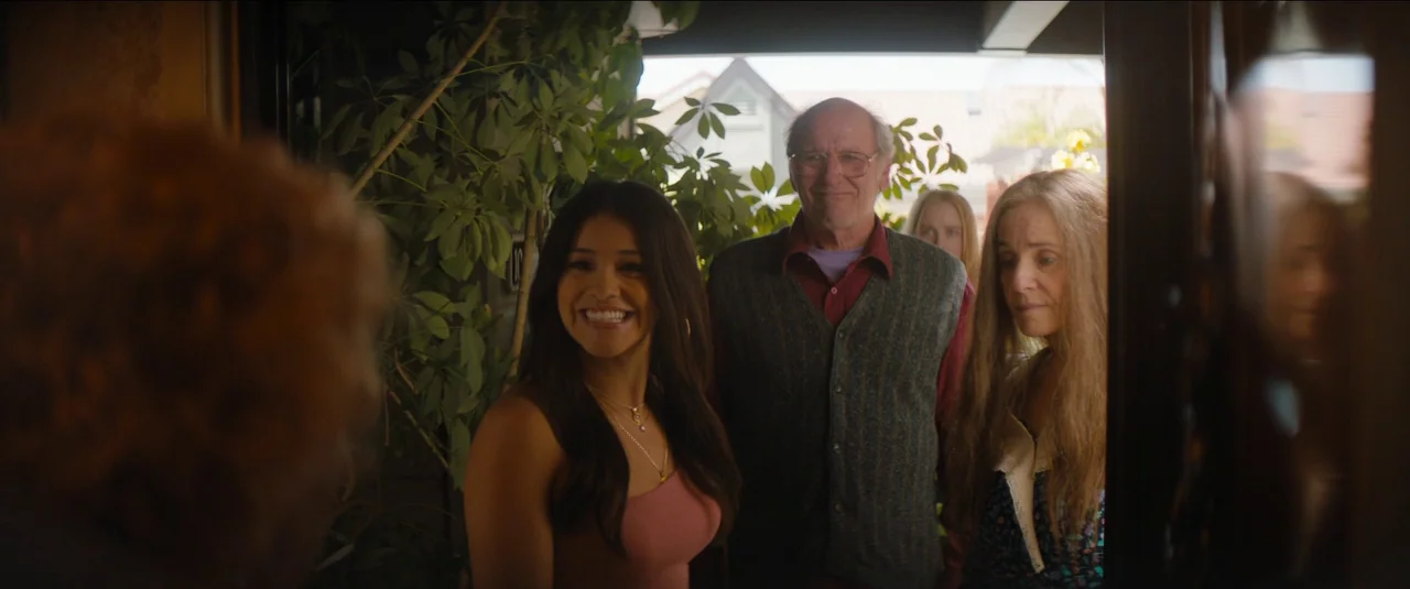 Kajillionaire (2020) movie still: medium — A group of three people stand on a suburban porch near dense green foliage. They smile toward a person…