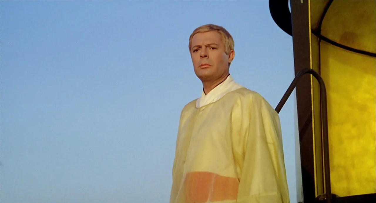 The 10th Victim (1965) movie still: medium — A man in a yellow tunic stands against a clear blue sky. He looks towards the left of the frame near a…