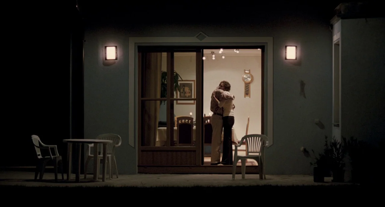 Revanche (2008) movie still: fullbody — A dark patio exterior faces a sliding glass door revealing a brightly lit dining area. A man and a woman…