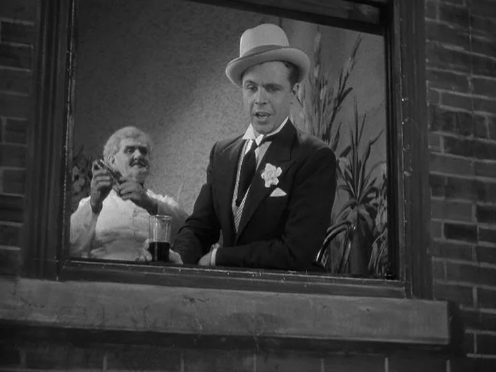 42nd Street (1933) movie still: medium — Two men stand inside an opening looking out through a brick building window. The foreground man in a hat…