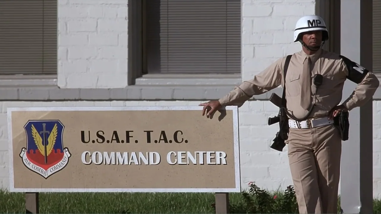 Crisis Point (2012) movie still: fullbody — A white brick exterior wall stands behind a tan command center sign. A military police officer in a tan…