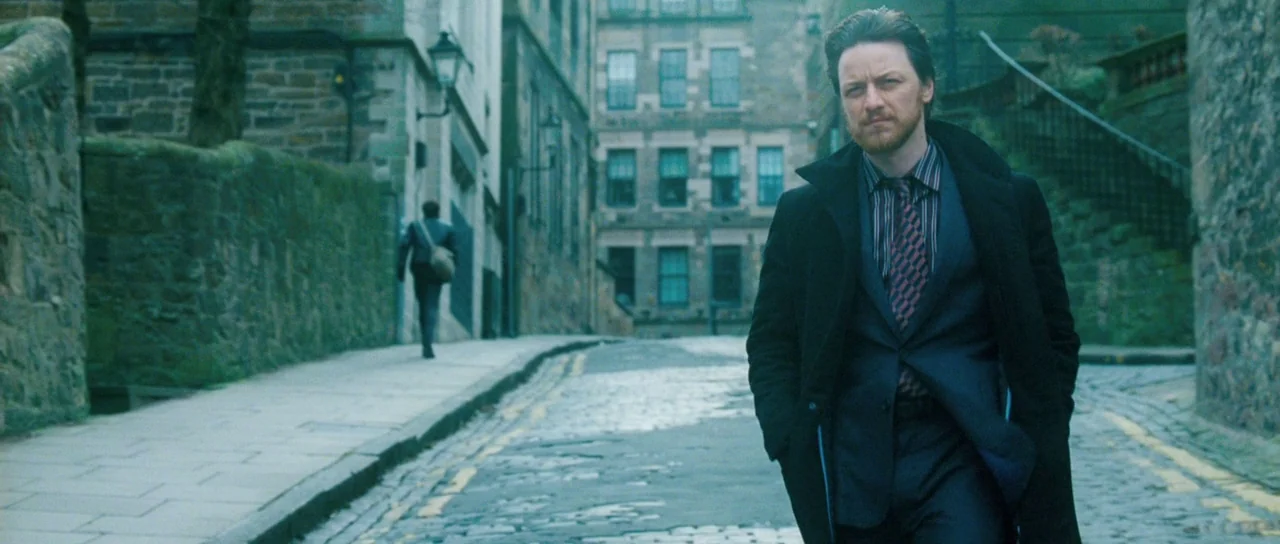 Filth (2013) movie still: medium — An old cobblestone street lined with tall stone buildings in a European city. A man in a dark coat walks toward…