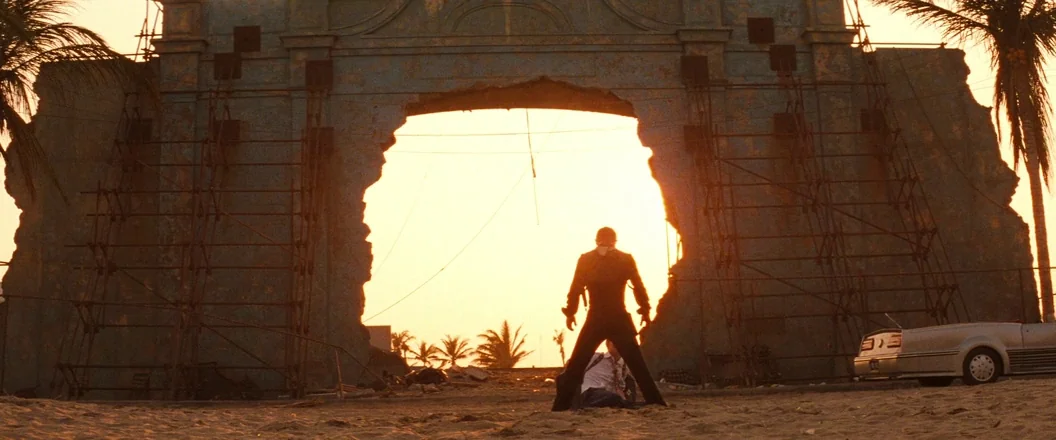 Romeo + Juliet (1996) movie still: wide — A massive, partially destroyed concrete archway stands on a sandy beach during sunset. A man stands in a…