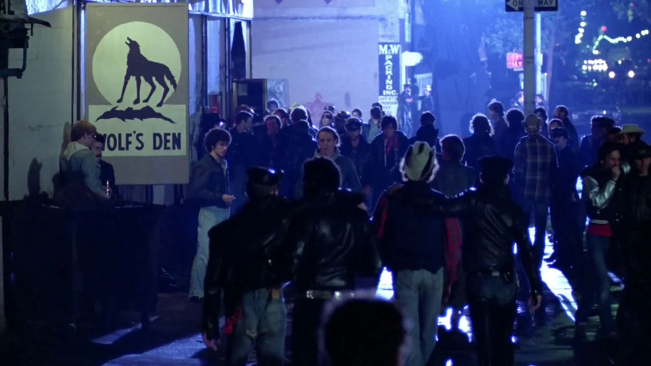 Cruising (1980) movie still: wide — A group of men walks through a dimly lit urban alleyway at night. A prominent sign depicting a howling wolf…