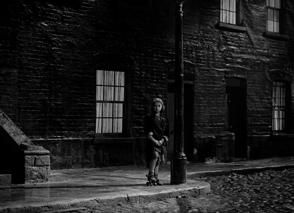 Odd Man Out (1947) movie still: fullbody — A dark city block features brick buildings with multiple windows at night. A young girl on roller skates…