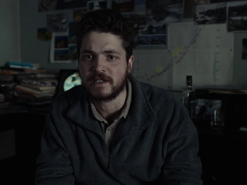 First Reformed (2017) movie still: medium — A cluttered room features a wall covered in various printed photographs and charts. A man sits in front…