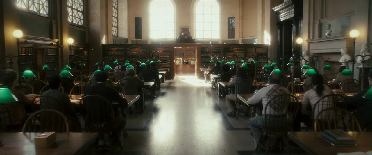 The Box (2009) movie still: wide — A quiet, grand room with rows of wooden study tables and green lamps. Many people are seated at the desks,…