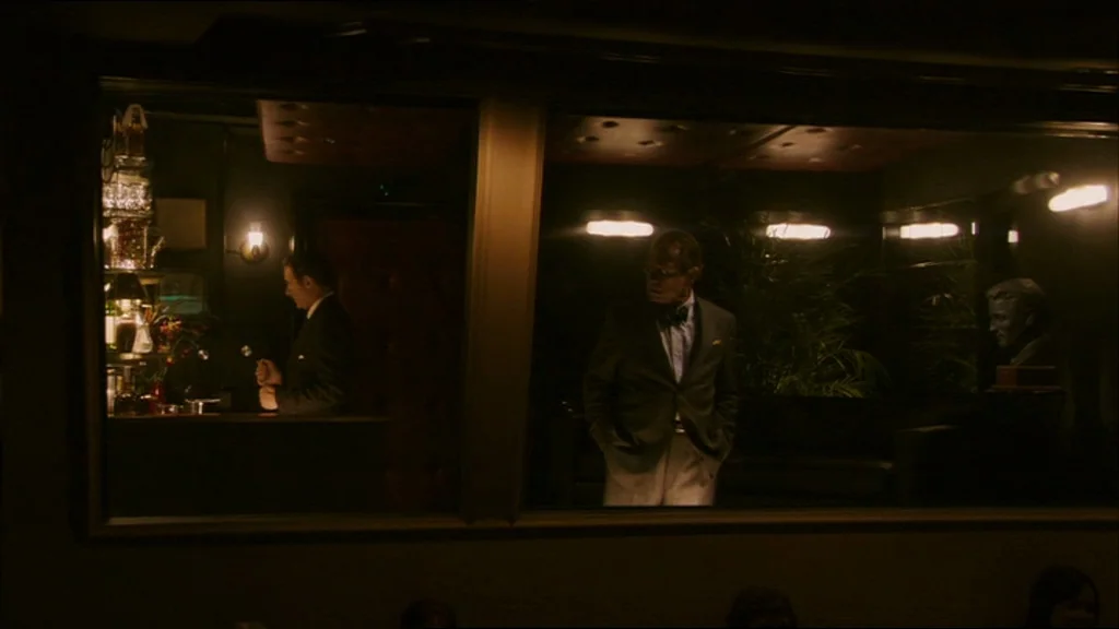 A Prairie Home Companion movie still: wide — A dimly lit club interior viewed through a large rectangular glass window. A bartender works near…