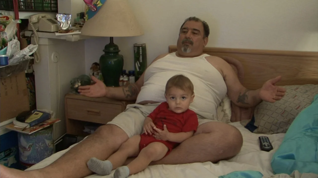 Homeless: The Motel Kids of Orange County (2010) movie still: medium — A cluttered bedroom with white walls and a large bed covered in linens. A man…