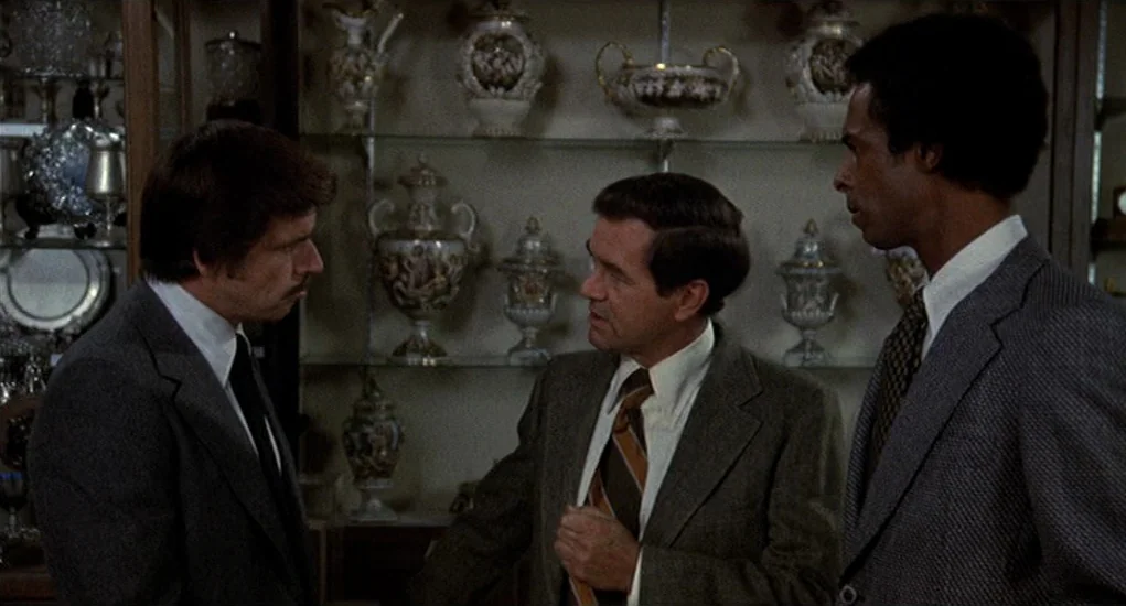 Family Plot (1976) movie still: medium — A well-lit interior of a shop containing shelves filled with antique ceramic vases. Three men in…