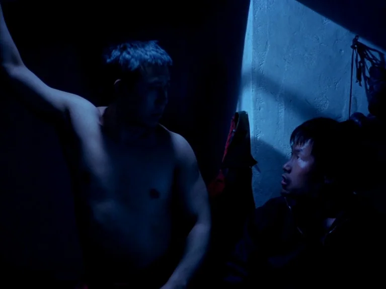 Black Snow (1990) movie still: medium — A dark, cramped room featuring a textured wall lit by a sharp sliver of light. Two men are positioned close…