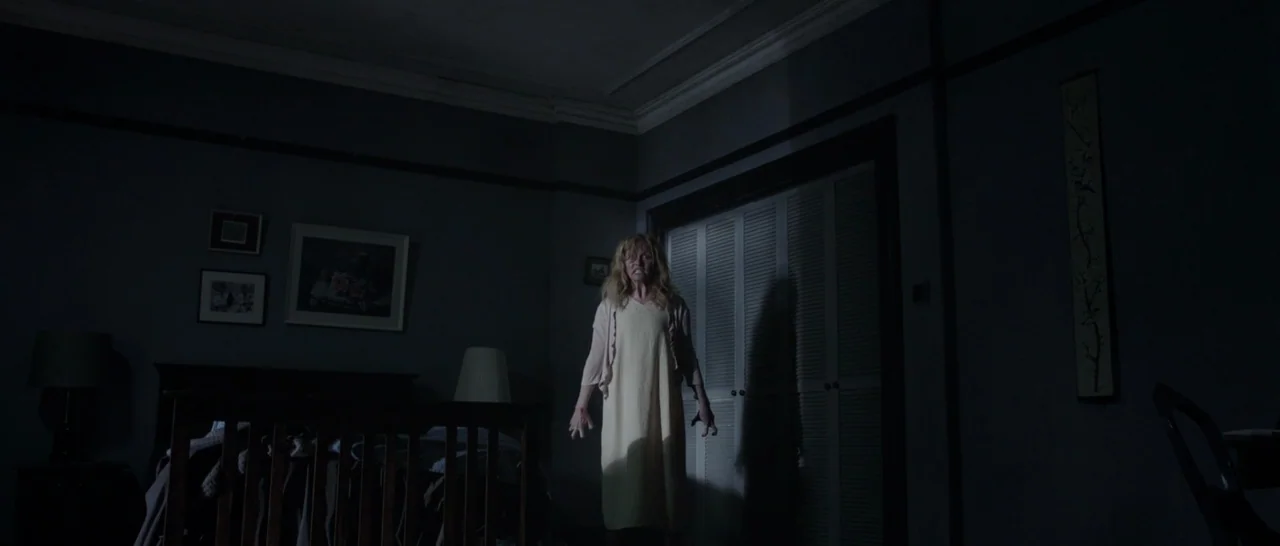The Babadook (2014) movie still: fullbody — A dark nursery contains a wooden crib, wall art, and shuttered closet doors. A woman stands in the…
