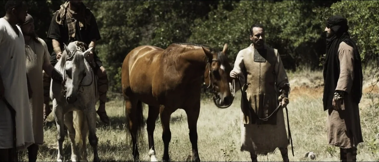 The Penitent Thief (2021) movie still: wide — A dry grassy field with dense green trees in the background under sunlight. Several men in historical…