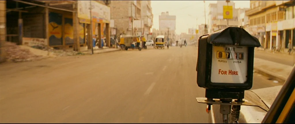 The Darjeeling Limited (2007) movie still: closeup — A taxi fare meter is mounted in the foreground of a sunny urban road. Distant buildings and a…
