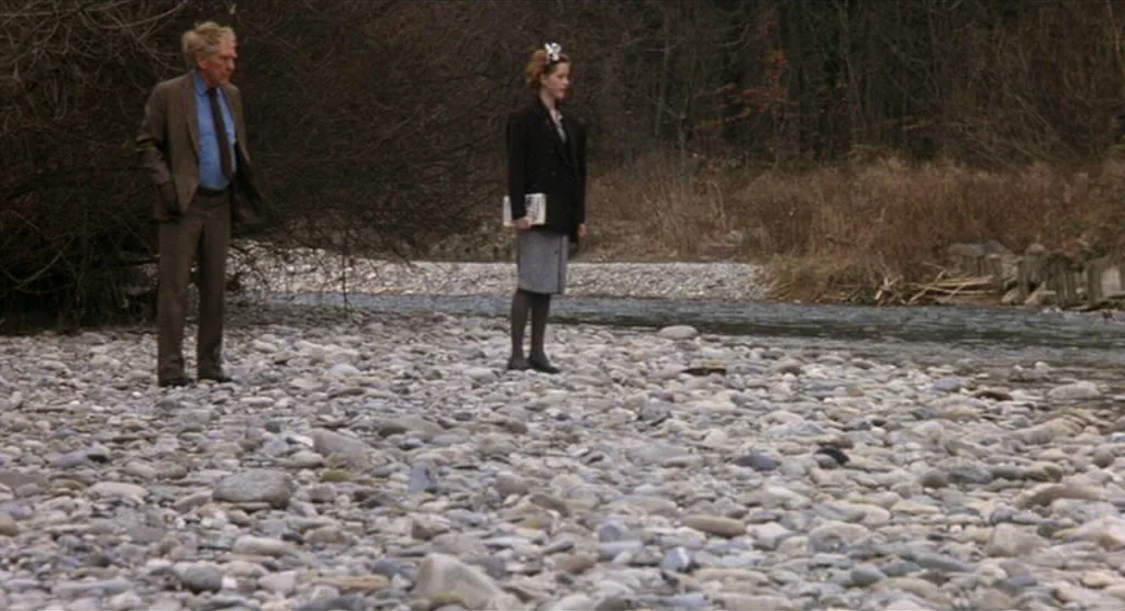 King Lear (1987) movie still: fullbody — A wide rocky riverbed borders a dense thicket of bare deciduous trees. An elderly man and a younger woman…
