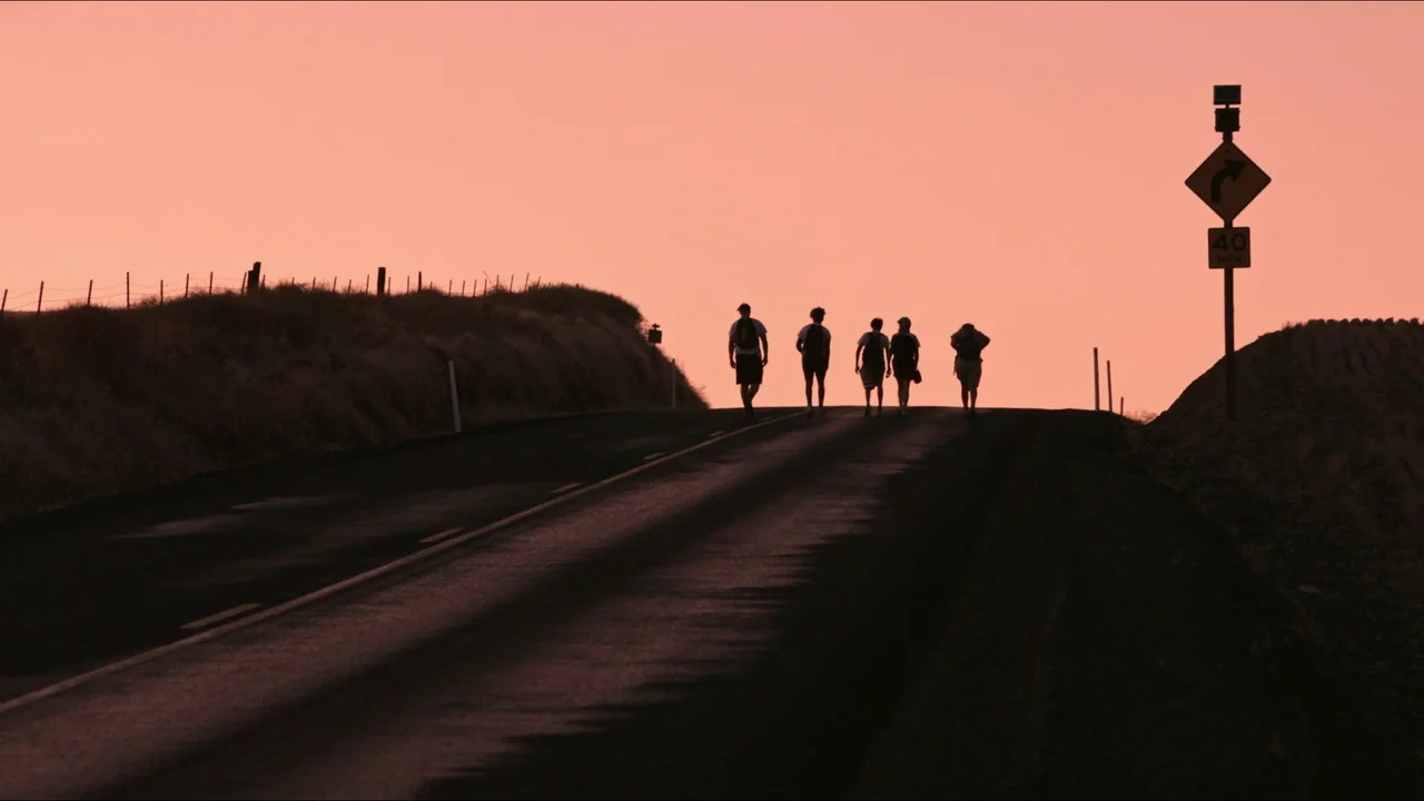 Gasoline Rainbow (2023) movie still: wide — An asphalt road rises toward a hill against a vibrant pink sunset sky. Five teenagers walk away from the…