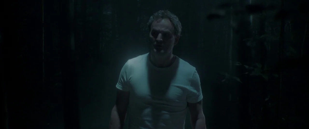 Pet Sematary (2019) movie still: medium — A man stands in a dimly lit, dark wooded area at night. He is illuminated by a soft overhead source…