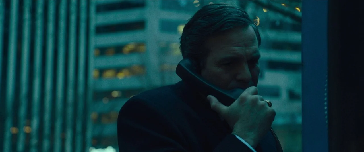 Dark Waters (2019) movie still: closeup — A man speaks on a telephone in an urban setting at night. Tall city buildings with illuminated windows are…