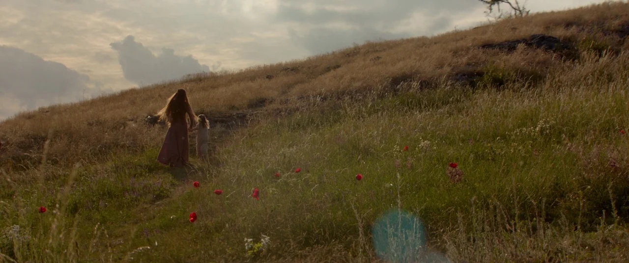 Ophelia (2018) movie still: wide — A wide, sloping hillside covered in dry golden grass and scattered red poppy flowers. An adult woman and a small…