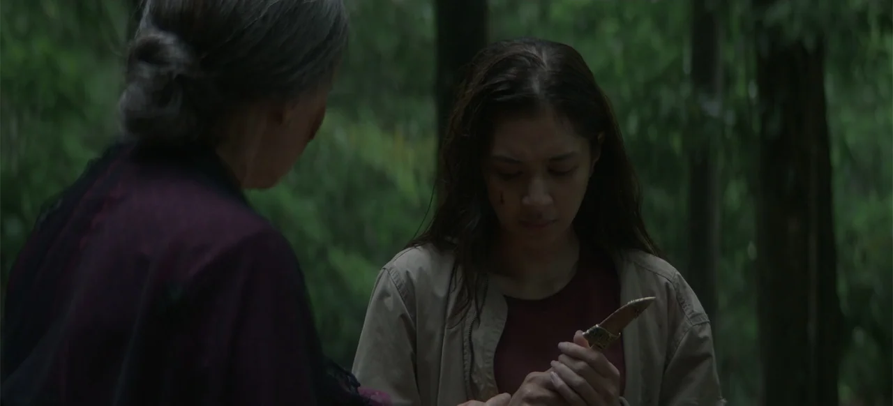A Thousand Days (2023) movie still: medium — An outdoor location surrounded by dense green trees and foliage in diffused daylight. A young woman…