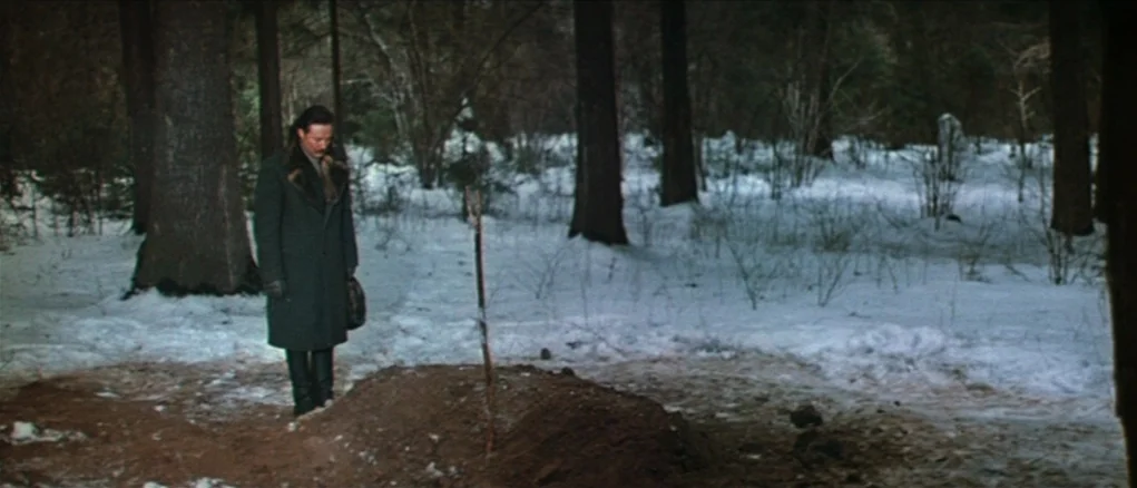 Dersu Uzala (1975) movie still: wide — A man stands alone in a snow-covered forest beside a fresh grave mound. Trees surround the clearing in the…