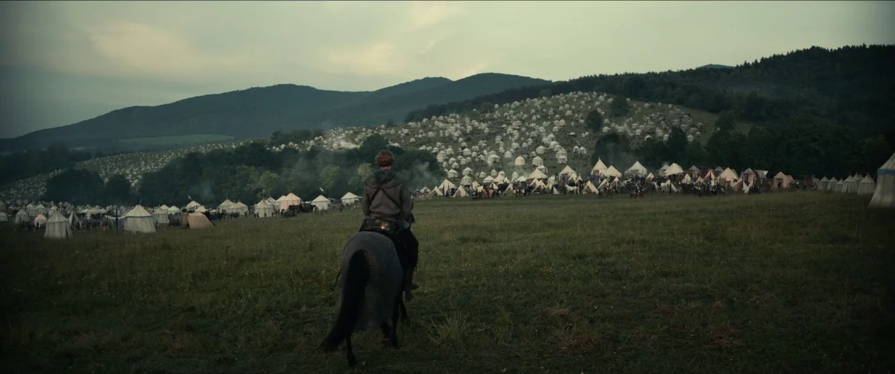 The King movie still: wide — A sprawling medieval military camp fills the hillside with numerous white conical canvas tents. A lone rider on a dark…