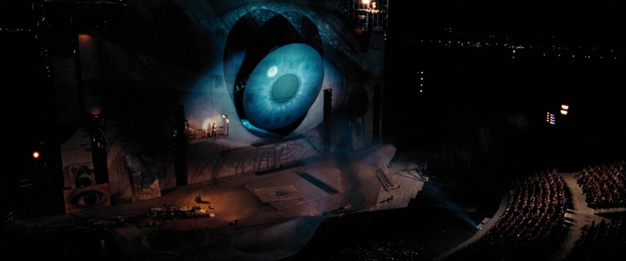 Quantum of Solace (2008) movie still: wide — An outdoor amphitheater stage features a massive illuminated projection of a blue human eye. Rows of…