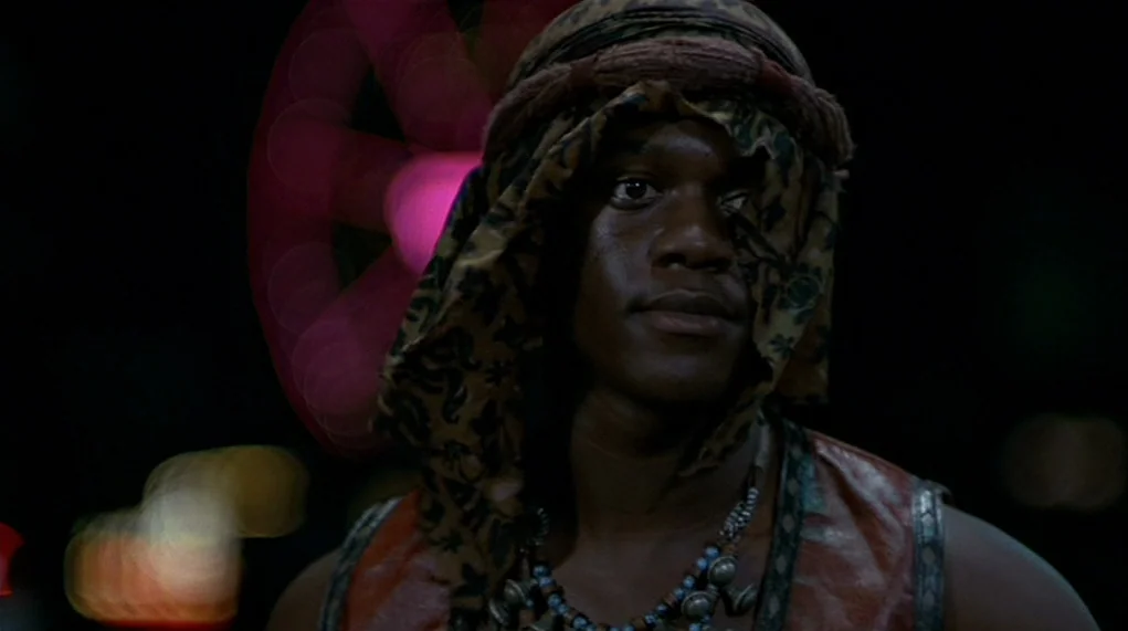 The Warriors (1979) movie still: closeup — A dark city street at night serves as the backdrop for a close portrait. A man wearing a headscarf looks…