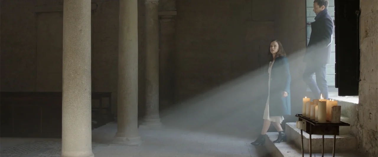 About Us (2017) movie still: medium — A stone-walled church interior features tall columns and a light-filled doorway leading outside. A woman in a…