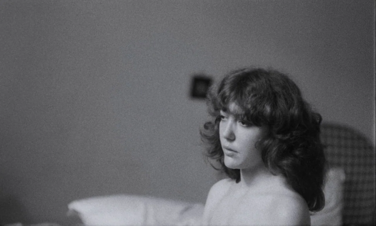 The Secret Son (1979) movie still: medium — A plain interior space features a person sitting in front of a white pillow. A young woman with wavy…