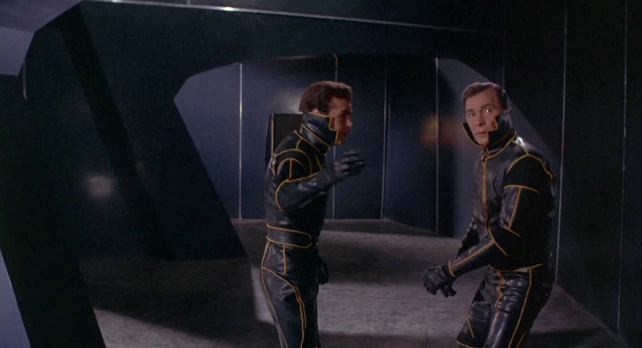 Planet of the Vampires (1965) movie still: medium — A dimly lit, windowless corridor features dark, panelled walls with sharp geometric angles. Two…