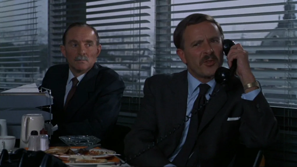 Frenzy (1972) movie still: medium — An office with venetian blinds overlooking a city skyline during a grey day. One man talks on a black telephone…
