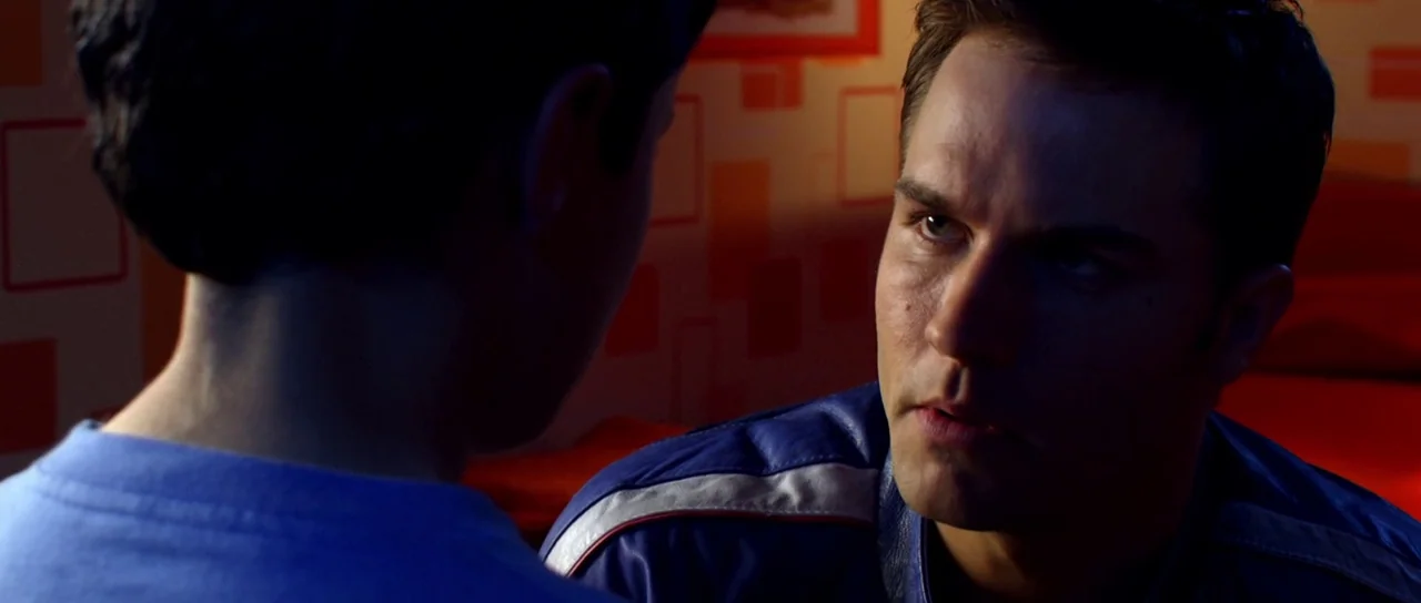Speed Racer (2008) movie still: medium — A dimly lit room with orange patterned wallpaper provides the backdrop for a conversation. A man wearing a…