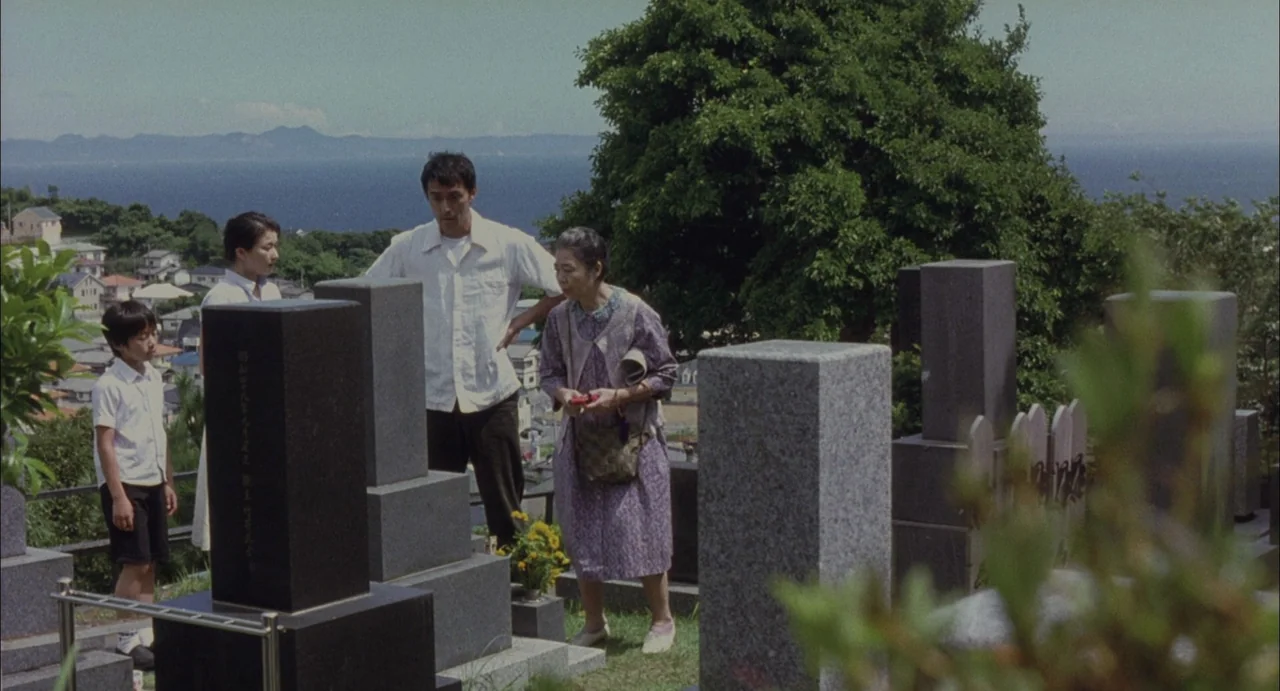 Still Walking (2008) movie still: wide — A Japanese hilltop cemetery overlooks a wide coastal bay with houses in the distance. Four people of…