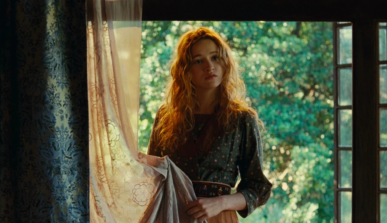 Renoir (2012) movie still: medium — A woman stands by an open window looking out at a densely wooded area. She holds a light sheer curtain back with…