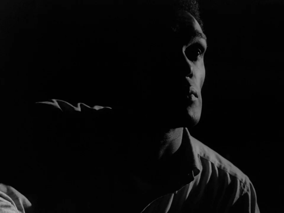Night of the Living Dead (1968) movie still: closeup — A man stands in an indeterminate dark space with deep shadows across his face. He is lit from…