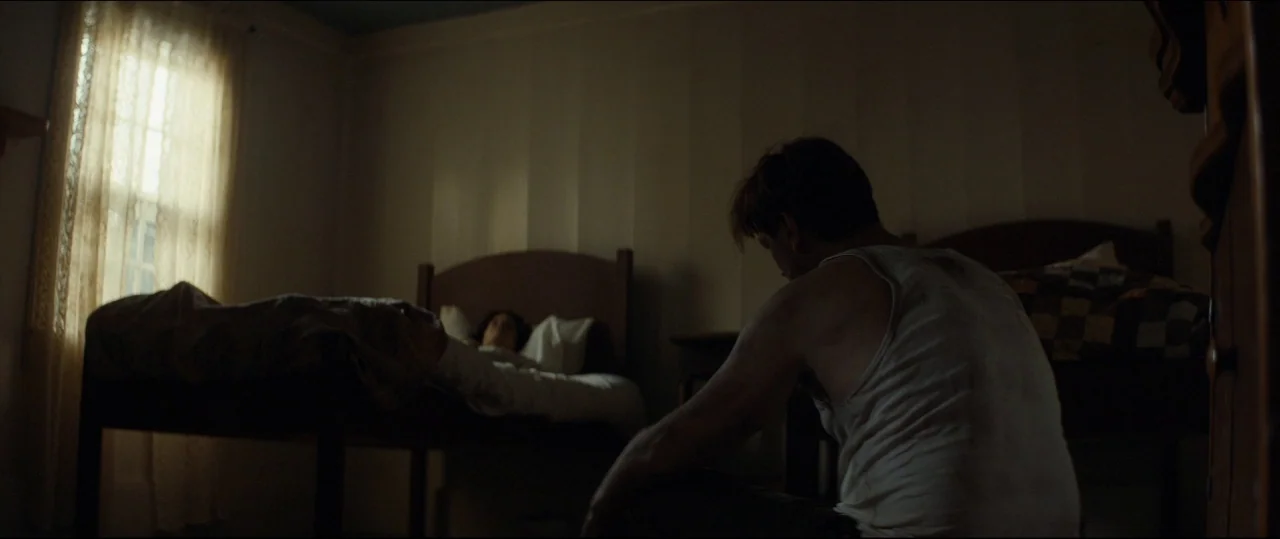 One & Two (2015) movie still: wide — A dimly lit room contains two wooden beds with simple linens. A man sits on the floor facing away while a woman…